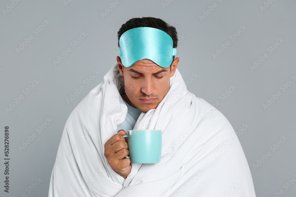 Man in sleeping mask wrapped with blanket holding cup on grey background Man in sleeping mask wrapped with blanket holding cup on grey background