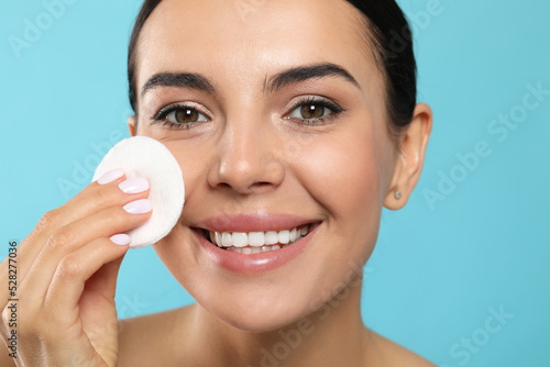 Young woman using cotton pad with micellar water on light blue background, closeup