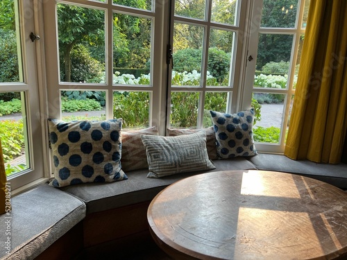 Wooden table near cozy bay window seat with cushions in room. Interior design photo