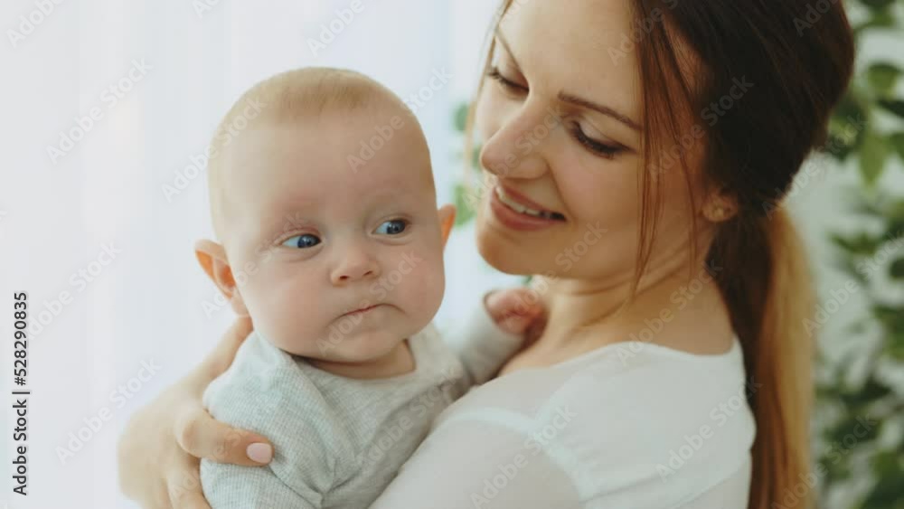 Close view of the caucasian mother carrying little baby at their new home. Portrait of the beautiful newborn baby that looking at the camera in mother hands. Motherhood concept Close view of the caucasian mother carrying little baby at their new home. Portrait of the beautiful newborn baby that looking at the camera in mother hands. Motherhood concept