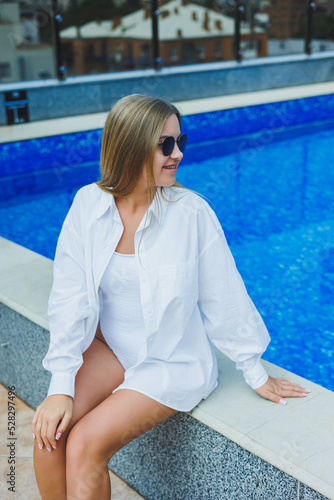 A cheerful young woman in a white swimsuit and a white shirt is sitting by the pool. A woman in sunglasses and a swimsuit. Summer vacation by the pool. Tanned girl