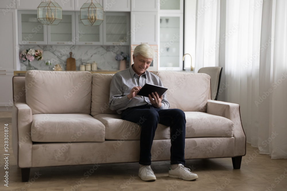 Cheerful engaged retired tablet user man sitting on home sofa, touching screen, using online app on gadget, shopping on Internet, watching media content, movie. Communication. Full length