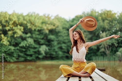 Hippie eco-activist woman traveler sits on a bridge by a lake with her arms outstretched with a hat and smiling sincerely © SHOTPRIME STUDIO
