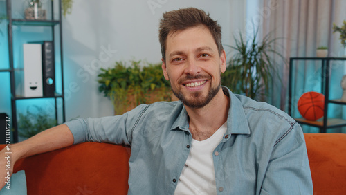 Close-up of happy calm adult man in shirt smiling friendly, glad expression looking away dreaming resting, relaxation feel satisfied concept good news. Young caucasian guy sitting on sofa at home room