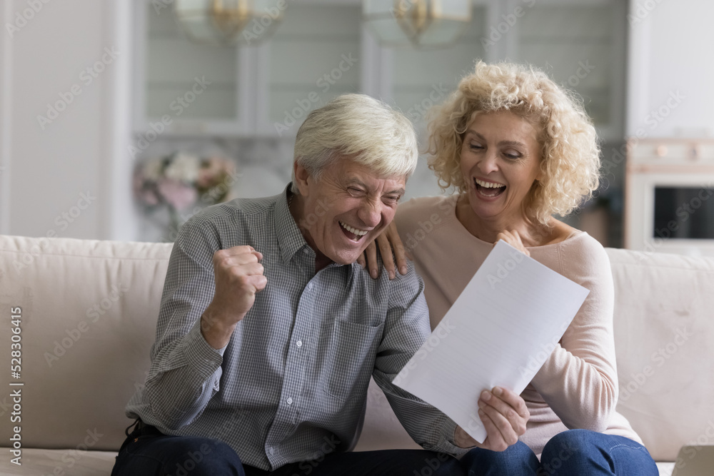 Fototapeta premium Happy excited senior retired couple reading paper document, making winner yes hand, celebrating success, achieve, luck, income, laughing, shouting for joy. Retirement, achievement