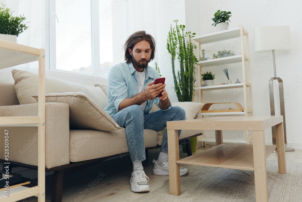 A man with a beard sits on the couch during the day and smiles at home and looks at his phone relaxing on his day off, life online on his phone texting with friends