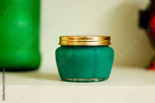 A jar with golden lid containing green wax or any green substance kept in a beige background with some green and red object in the background