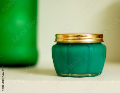 A jar with golden lid containing green wax or any green substance kept in a beige background with some green object in the background