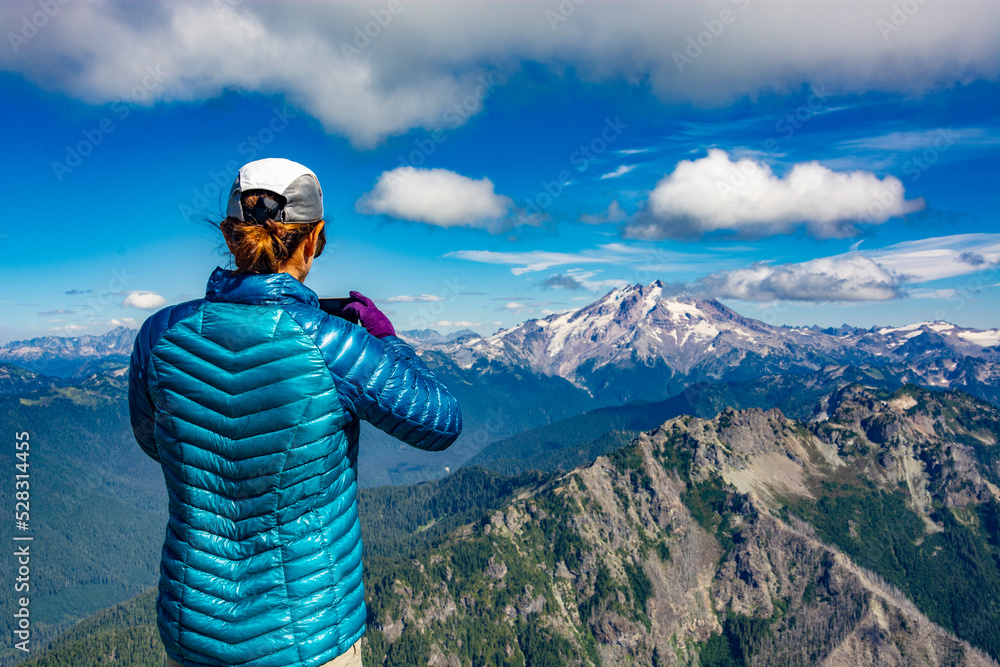Obraz premium Adventurous athletic female hiker taking a photo with her cellphone of jagged mountains in the in the Pacific Northwest.