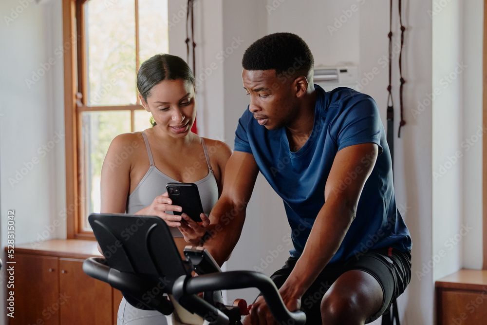 Obraz premium Two young multiracial friends using mobile phone on exercise bike at the gym