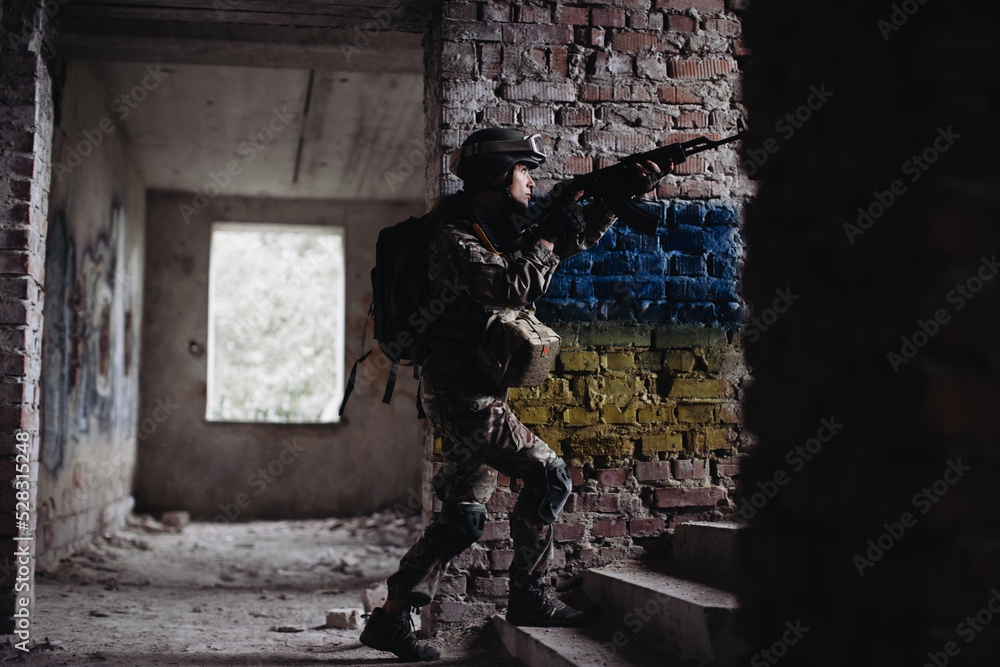 Fototapeta premium Military female soldier against the background of the flag of Ukraine. The flag of Ukraine is painted on a brick wall, a Ukrainian defender with a weapon in his hands.
