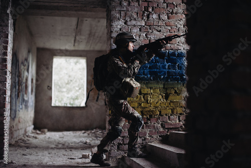 Military female soldier against the background of the flag of Ukraine. The flag of Ukraine is painted on a brick wall, a Ukrainian defender with a weapon in his hands.