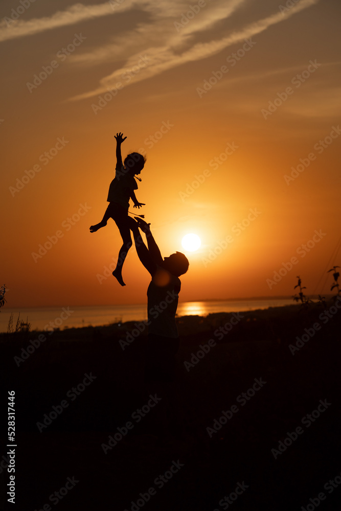 Silhouette of a man and a child against the sunset.Dad raises his son against the orange sky.People are having fun at sunset.The silhouette of a happy family standing against the sunset