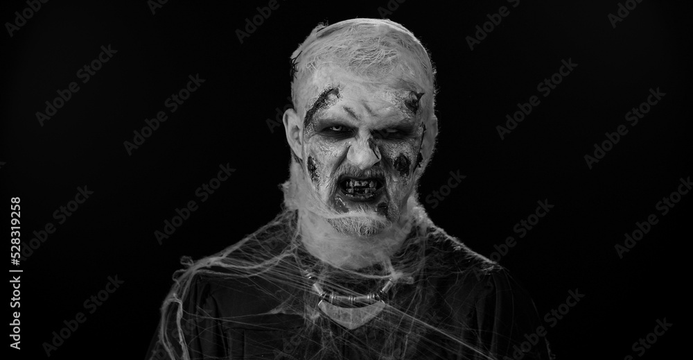 Fototapeta premium Sinister man with horrible scary Halloween zombie makeup in convulsions making faces, looking ominous, trying to scare. Dead guy with wounded bloody scars face isolated against black wall background