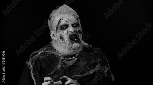 Scary man with horrible scary Halloween zombie makeup in convulsions making faces, looking ominous, trying to scare. Dead guy with wounded bloody scars face isolated against black wall background