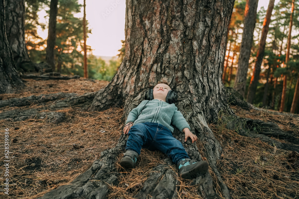 A little boy, a child, a toddler lies on the ground in the forest by a tree and listens to music through large headphones.