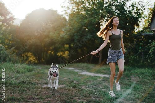 A woman runs with her dog in the summer in a park among greenery in nature outside the house. Lifestyle in sports and walking with a husky dog ​​on a leash