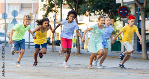Cheerful active tween boys and girls in colorful clothes running holding hands along summer city street..