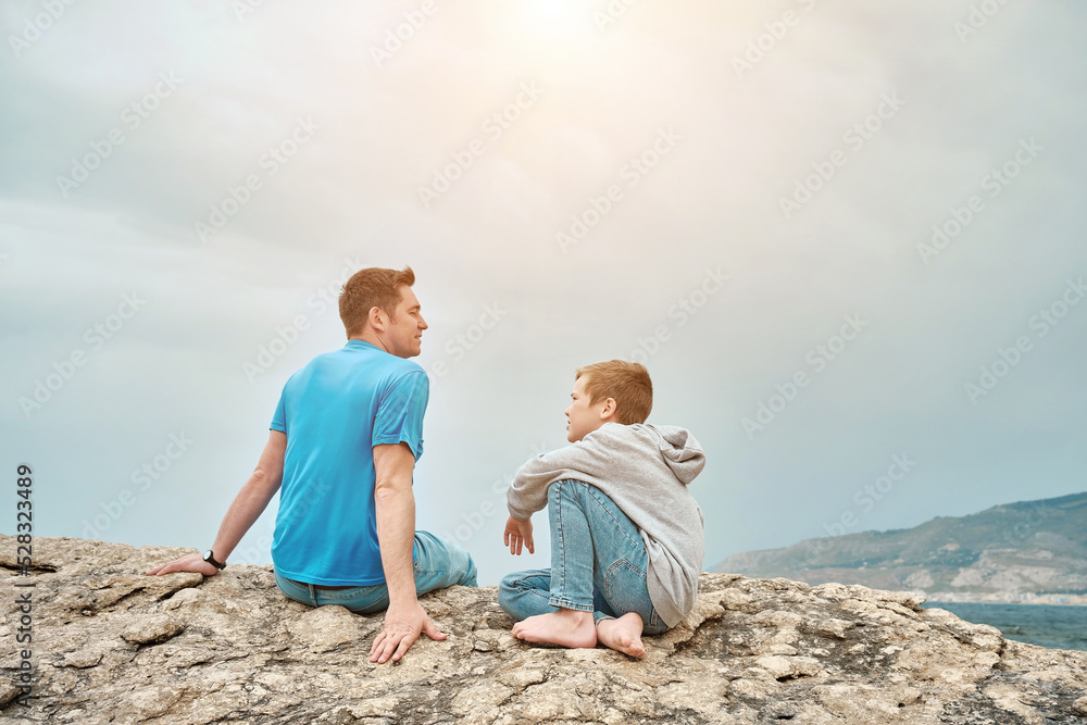 Happy family, father and son bonding, sitting on stone by the sea looking at view enjoying summer vacation. Togetherness Friendly concept
