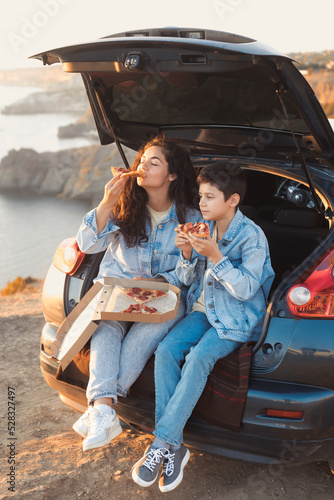Full body mother and teen son sitting in the trunk of a car and eating slices of take away pizza from box. Family vacation trip © Evgeniya Grande