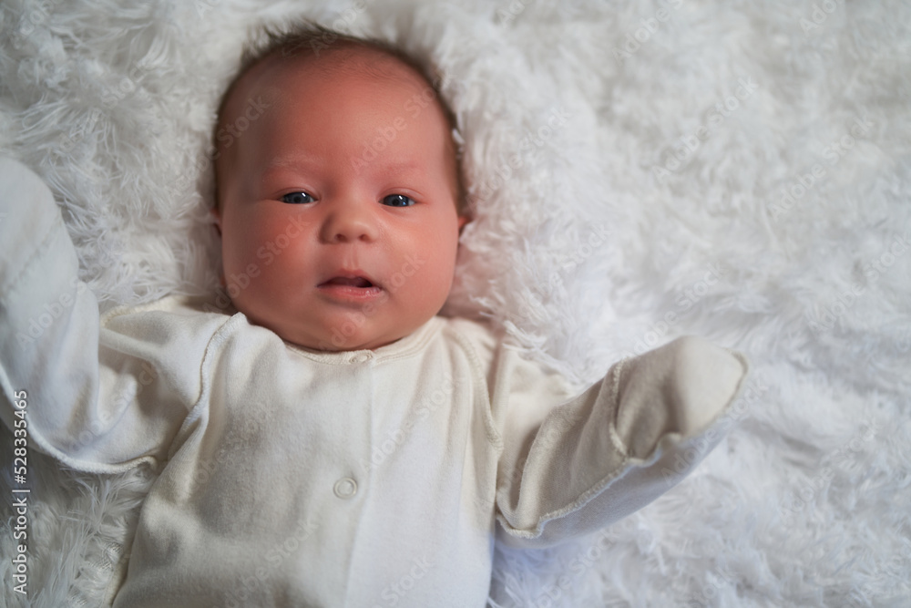 A newborn baby in white clothes lies on a feather blanket looking at the camera A newborn baby in white clothes lies on a feather blanket looking at the camera