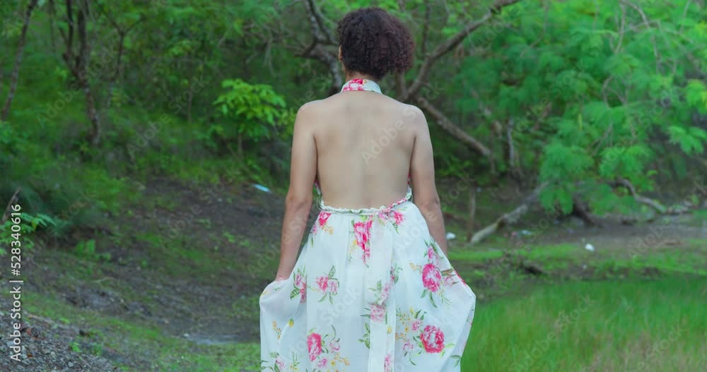A young woman walking in the shaded part of a park on a sunny day wearing a long flower dress A young woman walking in the shaded part of a park on a sunny day wearing a long flower dress