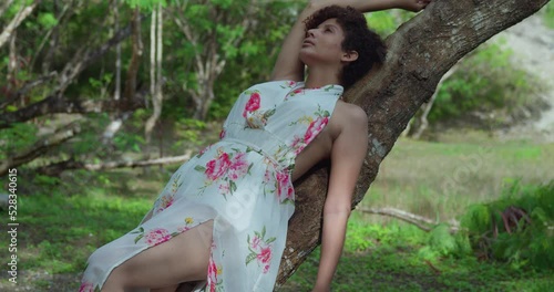 Curly short hair latina girl in a long dress lays on a tree log in a park on a sunny day photo