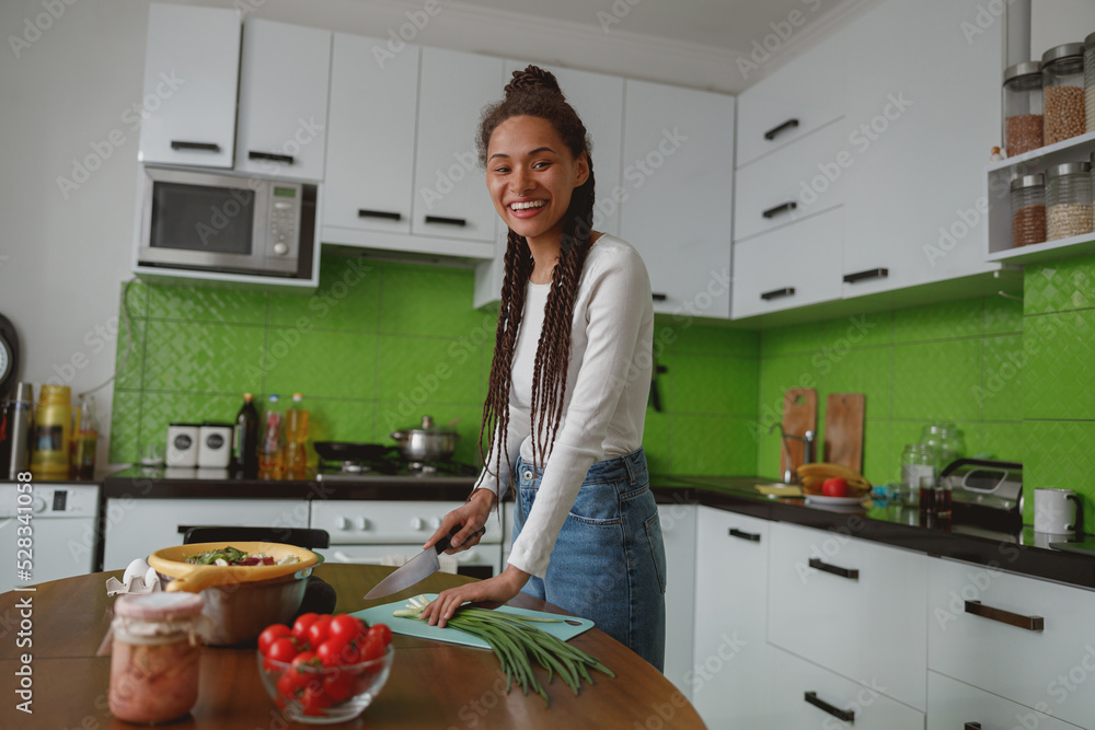Joyful positive mixed-race woman cooking in kitchen indoor preparing fresh salad, healthy food