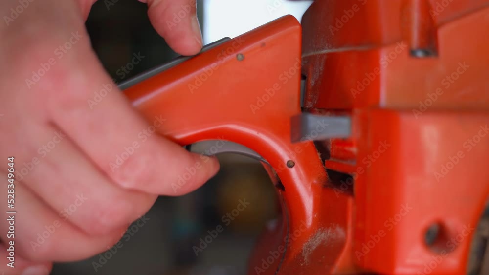 The hand presses the accelerator button on the handle of an orange chainsaw close-up. Maintaining the operating speed of the engine, when warming up to operating temperature The hand presses the accelerator button on the handle of an orange chainsaw close-up. Maintaining the operating speed of the engine, when warming up to operating temperature