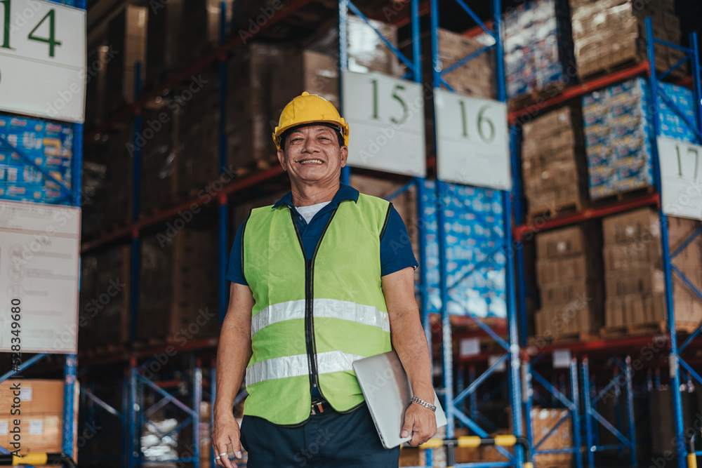Warehouse Workers standing and holding Laptop in Logistic center. Asian senior male worker wearing safety vest and Hard Hat to working about shipment in storehouse, Storage Distribution Center. Warehouse Workers standing and holding Laptop in Logistic center. Asian senior male worker wearing safety vest and Hard Hat to working about shipment in storehouse, Storage Distribution Center.