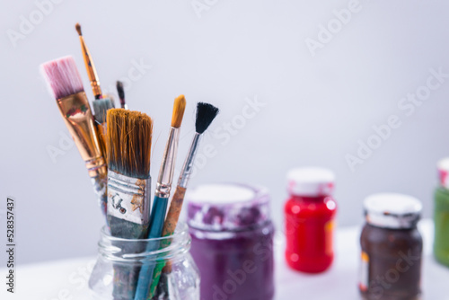 High quality photography. Close up to a glass beaker filled with an artist's brushes and paint pots in the background in a white room. Artistic theme.