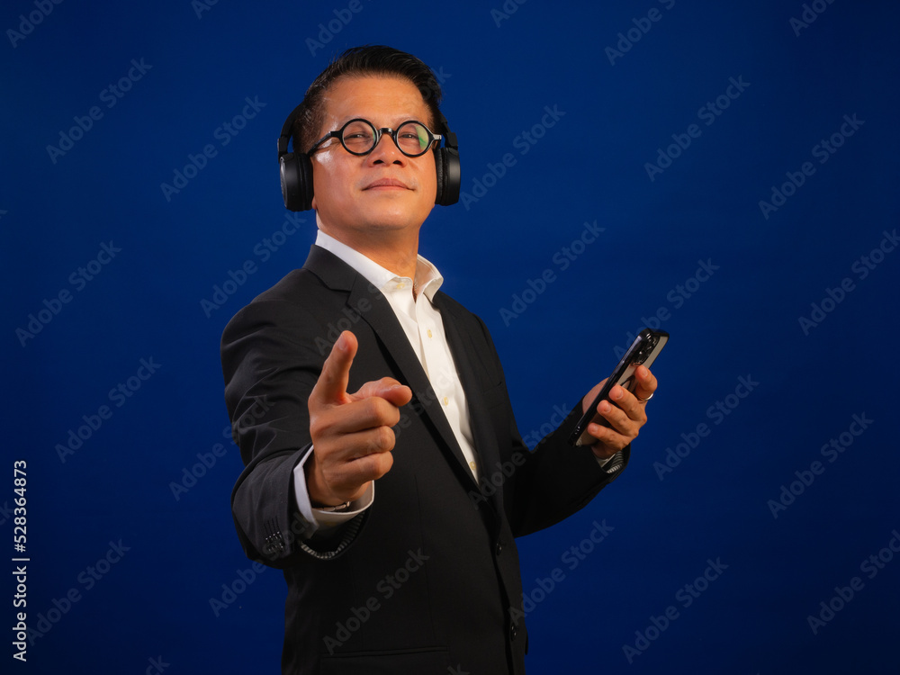 Portrait of smart middle-aged asian confident successful businessman wearing suit listening music with wireless headphones and smartphone on blue background in studio, looking happy leadership Portrait of smart middle-aged asian confident successful businessman wearing suit listening music with wireless headphones and smartphone on blue background in studio, looking happy leadership