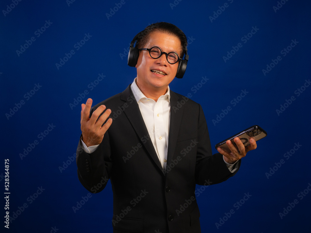 Portrait of smart middle-aged asian confident successful businessman wearing suit listening music with wireless headphones and smartphone on blue background in studio, looking happy leadership Portrait of smart middle-aged asian confident successful businessman wearing suit listening music with wireless headphones and smartphone on blue background in studio, looking happy leadership