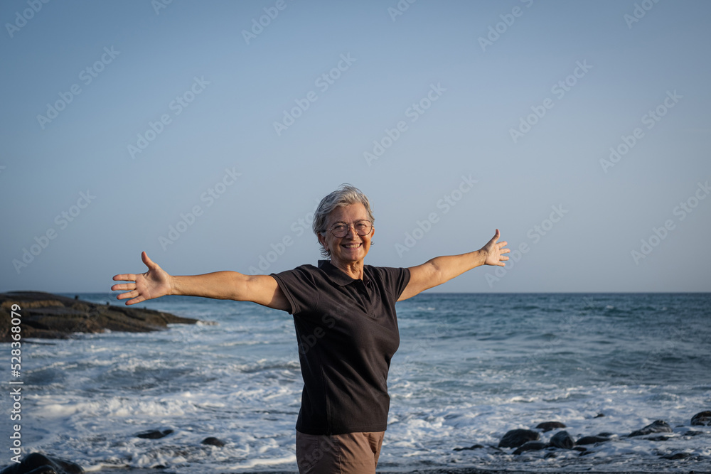 Happy caucasian senior woman standing on the beach at sunset with outstretched arms - elderly female enjoying free time, vacation or retirement