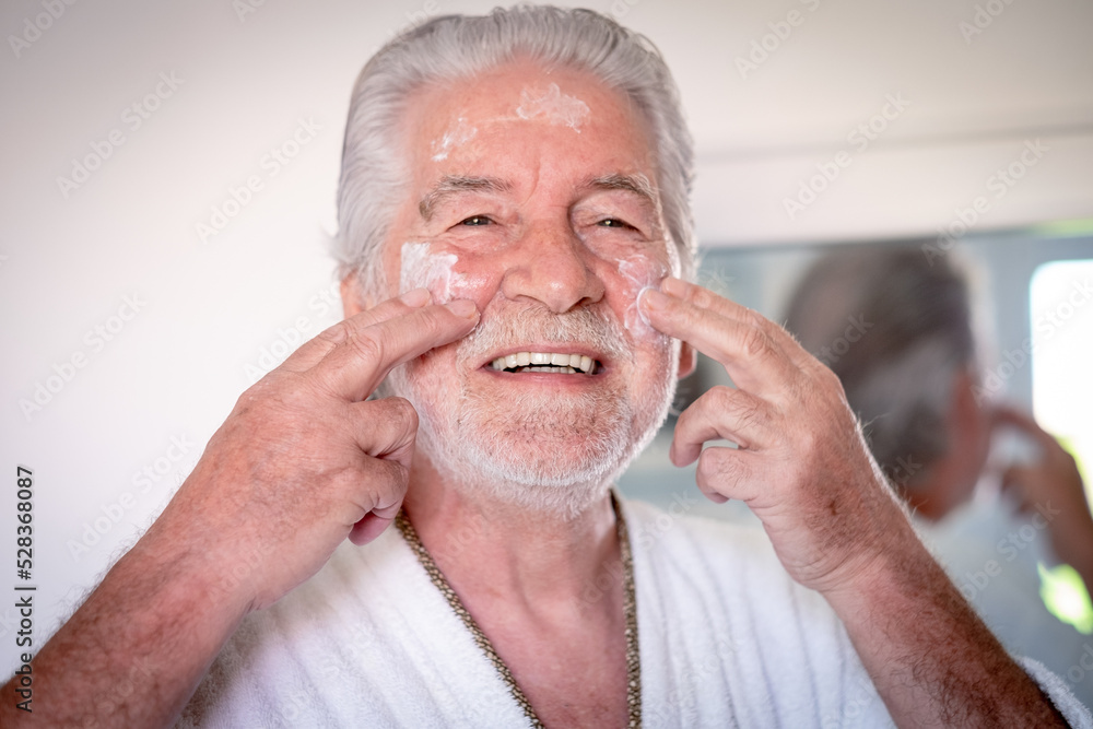 Obraz premium Skin care. Handsome elderly bearded man in bathrobe applying cream on his face as a daily routine