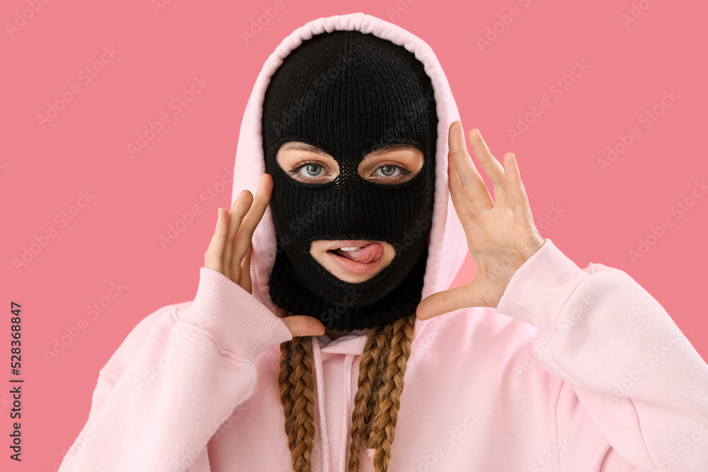 Young woman in balaclava with hands near face showing tongue on pink background