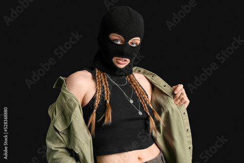 Young woman in balaclava touching shirt on black background