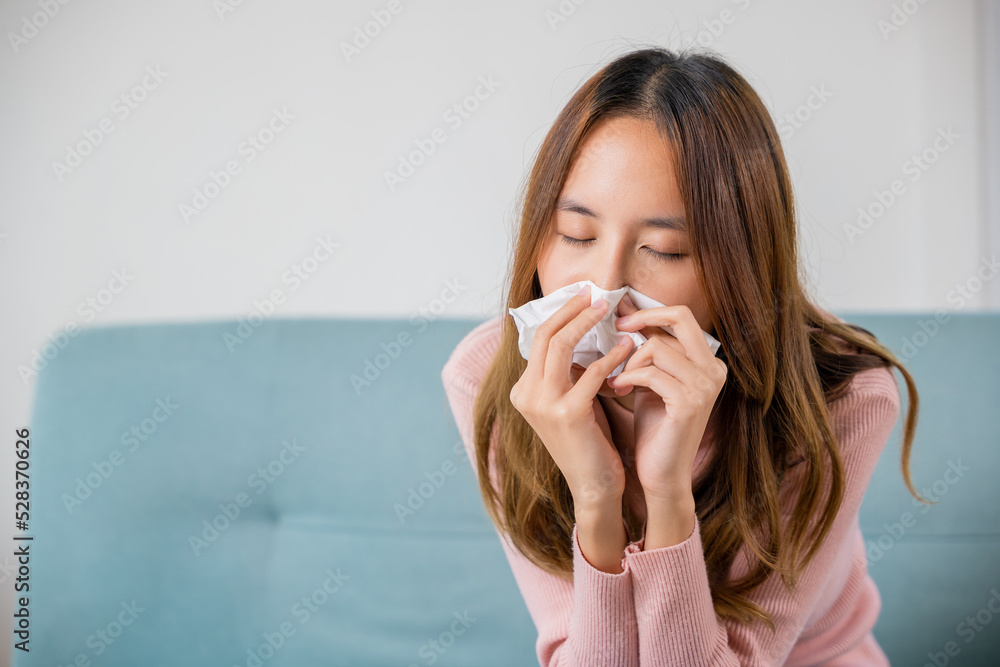 Ill Asian young woman cold covered with blanket sitting on sofa and sneeze with tissue paper at home, Female sick allergic blowing nose sneezing in tissue at home, Flu health care Ill Asian young woman cold covered with blanket sitting on sofa and sneeze with tissue paper at home, Female sick allergic blowing nose sneezing in tissue at home, Flu health care