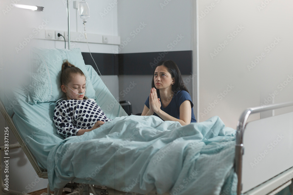 Fototapeta premium Ill kid under treatment sleeping in hospital pediatrics ward room while worried mother sitting next to her. Sick little girl resting on patient bed while mother praying beside her