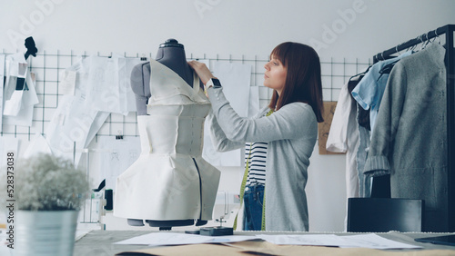 Young attractive seamstress is carefully measuring tailor's dummy with tape to make new garment with these measurements. Busy working day in clothing workshop concept.