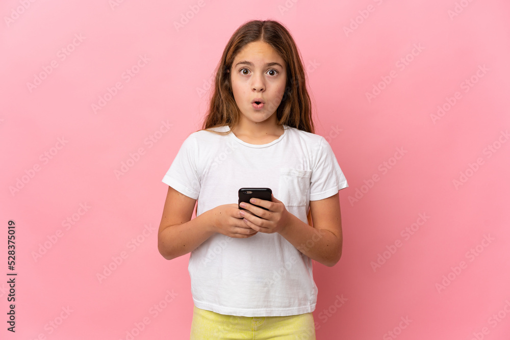 Child over isolated pink background looking at the camera while using the mobile with surprised expression Child over isolated pink background looking at the camera while using the mobile with surprised expression