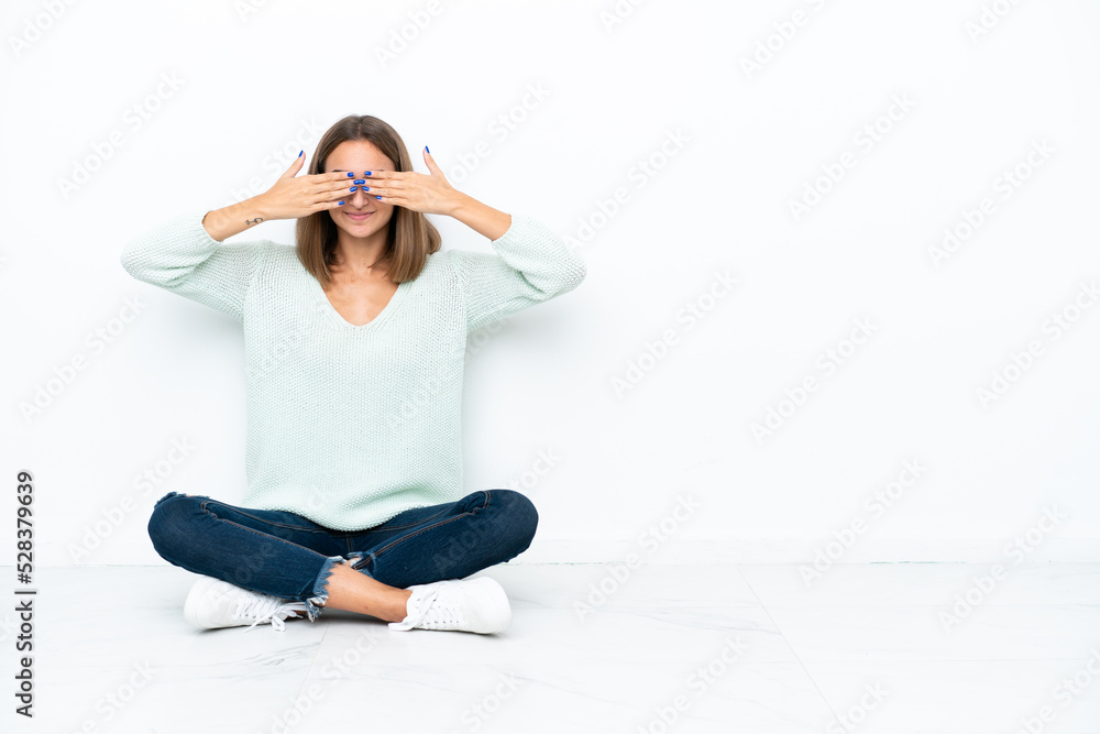 Fototapeta premium Young caucasian woman sitting on the floor isolated on white background covering eyes by hands