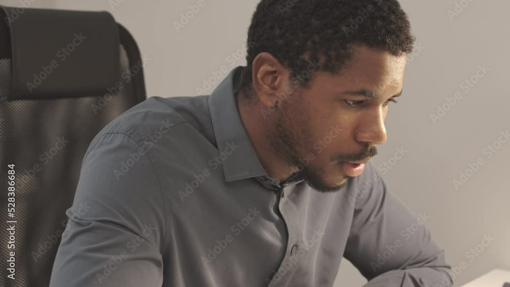Chest up slowmo of concentrated young Black man in dark blue shirt overworking late at night sitting by office desk Chest up slowmo of concentrated young Black man in dark blue shirt overworking late at night sitting by office desk