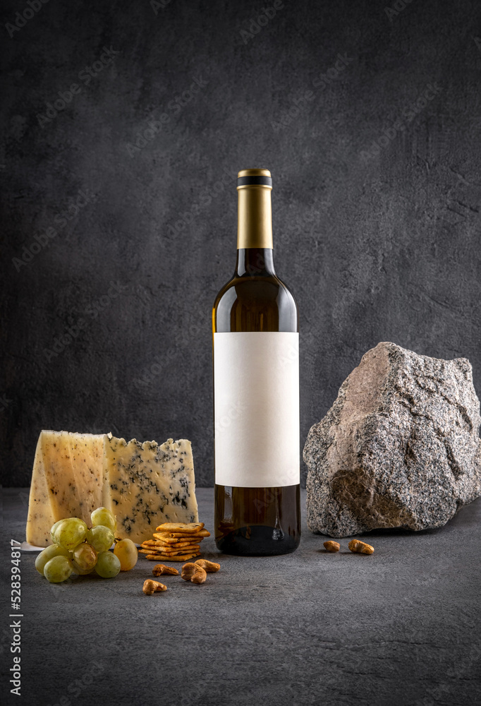 Naklejka premium Glass and bottle of white wine with grapes on dark background. White wine with cheese and fruits. Wine bottle mockup with blank labels to place your design. Space for text