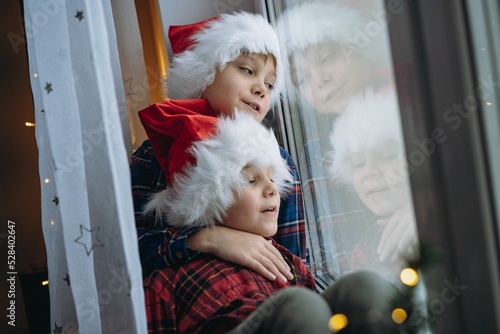 cute caucasian children wearing snta hats and plaid shirts sitting hugging by the window waitinf for christmas photo