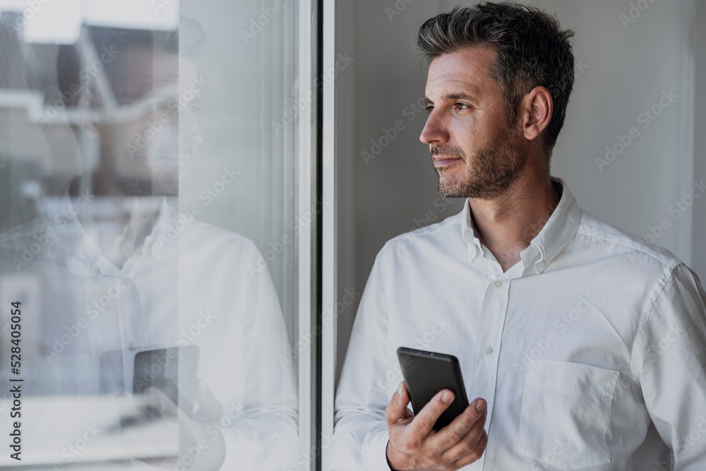 Thoughtful businessman holding mobile phone looking through window Thoughtful businessman holding mobile phone looking through window