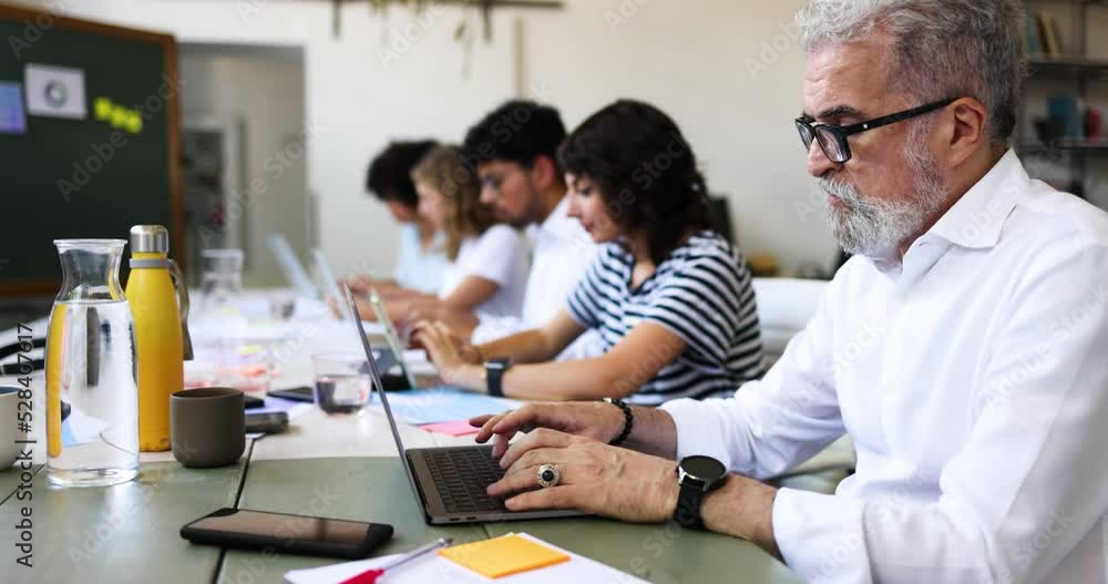 Senior businessman sitting at table with colleagues working on laptop Senior businessman sitting at table with colleagues working on laptop