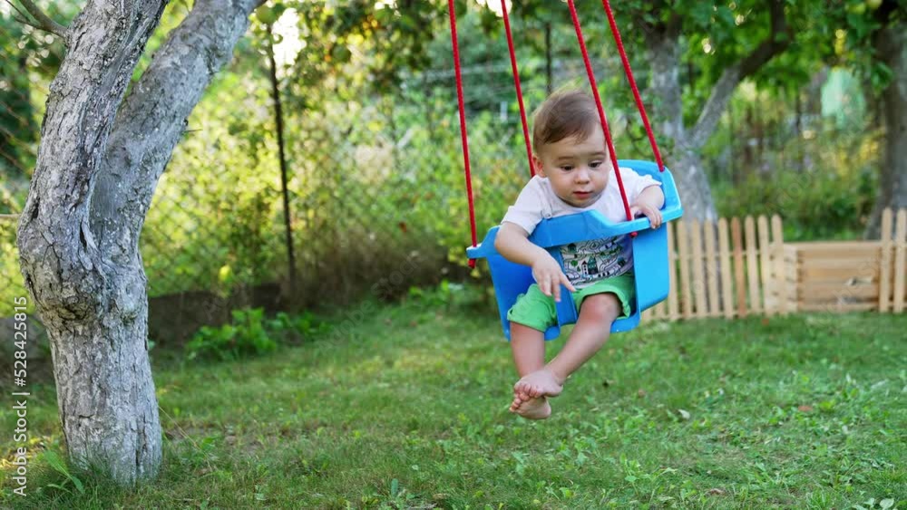 Lovely barefoot Caucasian boy swinging in a blue swing hanging from a tree. Toddler waving hand happily swaying. Garden background. Lovely barefoot Caucasian boy swinging in a blue swing hanging from a tree. Toddler waving hand happily swaying. Garden background.