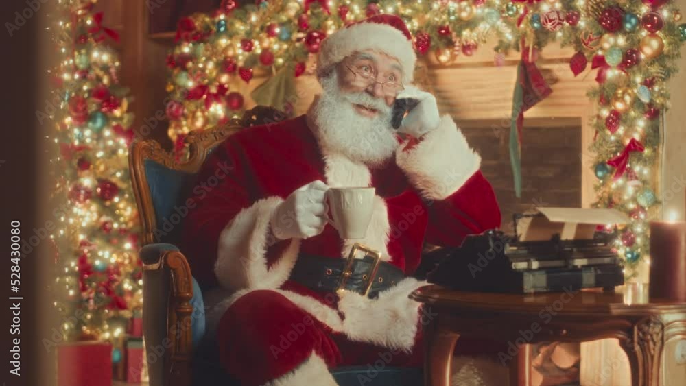 Medium slowmo of big bearded red Santa drinking hot tea from mug and talking on mobile phone while sitting in his char by typewriter in festively decorated room with fireplace Medium slowmo of big bearded red Santa drinking hot tea from mug and talking on mobile phone while sitting in his char by typewriter in festively decorated room with fireplace