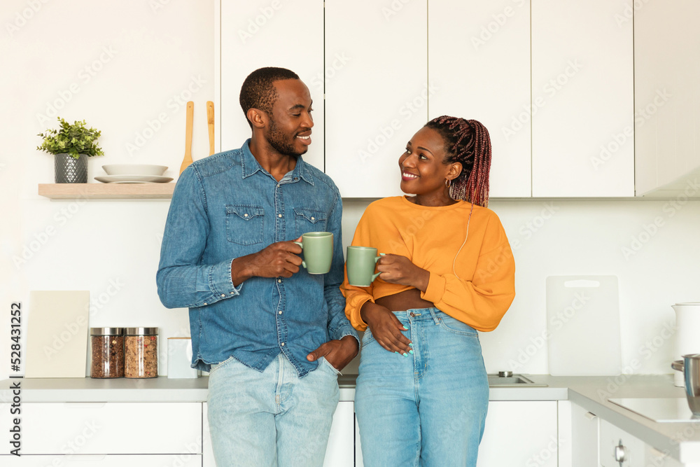 Fototapeta premium Happy african american spouses drinking morning coffee and talking, standing in kitchen interior, copy space